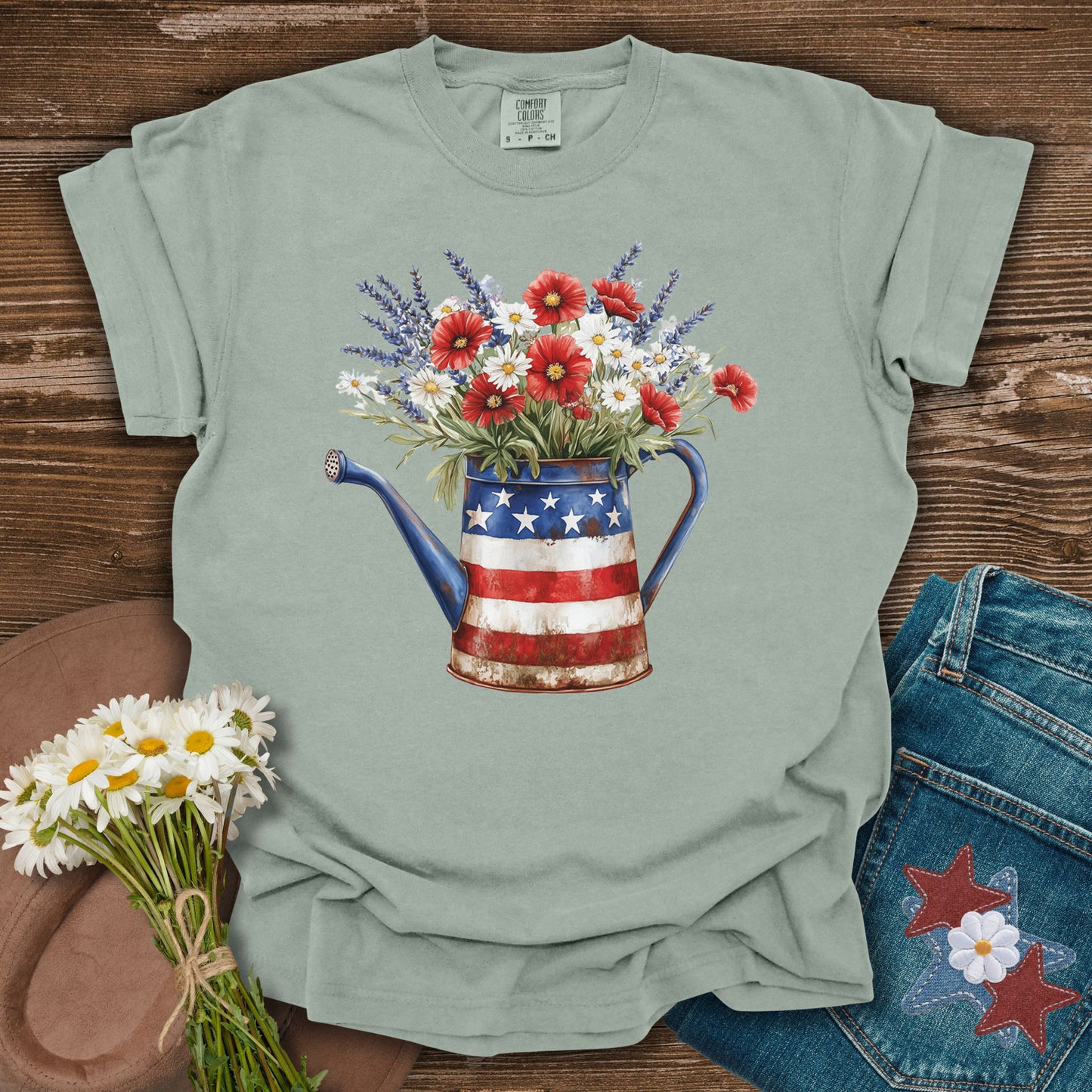 Garment-dyed t-shirt with a rustic patriotic watering can overflowing with lavender, daisies, and red poppy-like flowers.