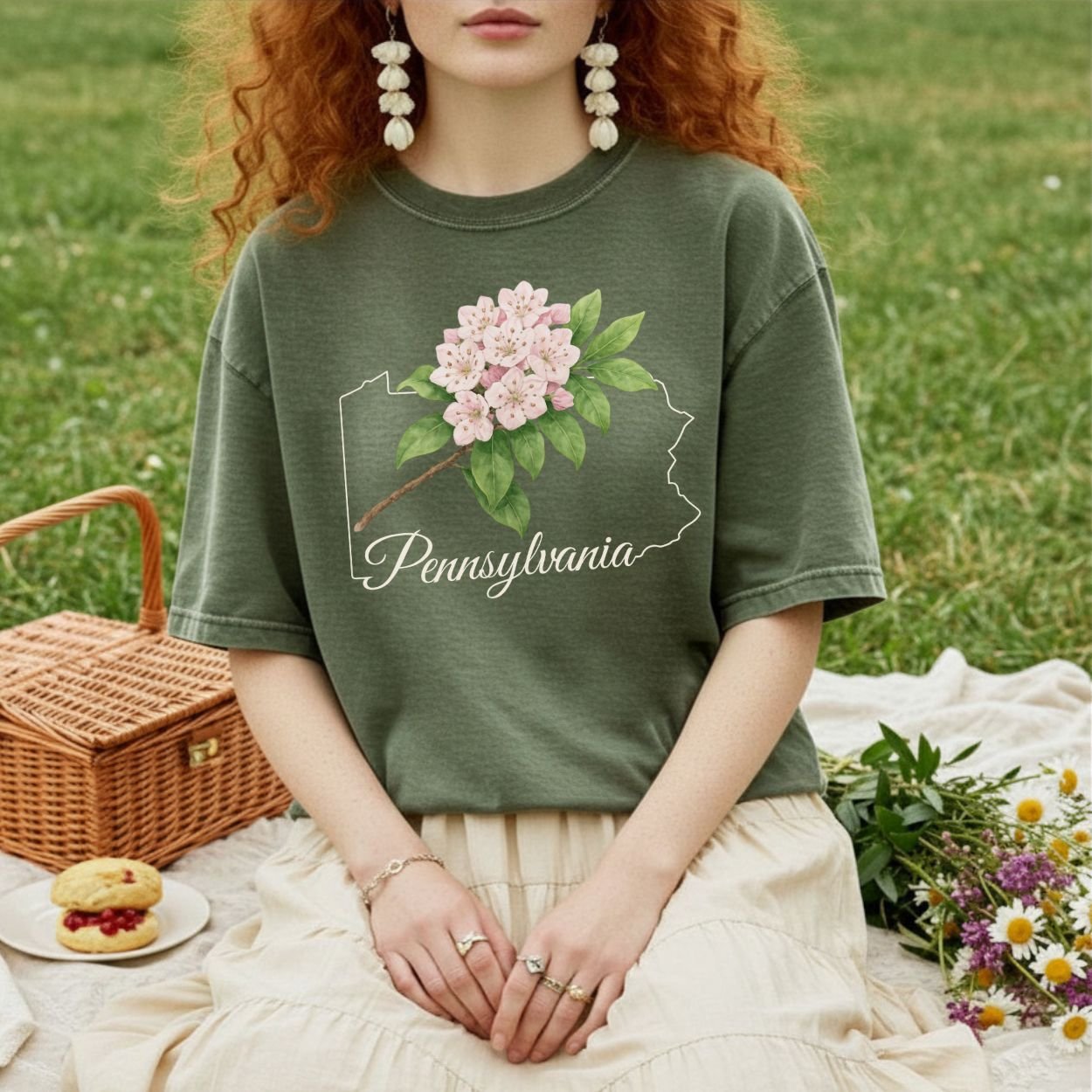 Garment-dyed olive green t-shirt with pink mountain laurel over a Pennsylvania outline, worn by a person sitting on a picnic blanket.