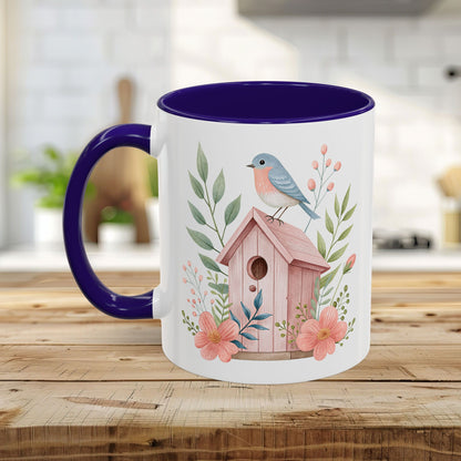 White ceramic mug with a pink birdhouse, bluebird, and peach blossoms, featuring a purple handle and interior.