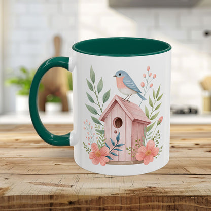 White ceramic mug with a green handle and interior, featuring a watercolor pink birdhouse, bluebird, and peach blossoms.