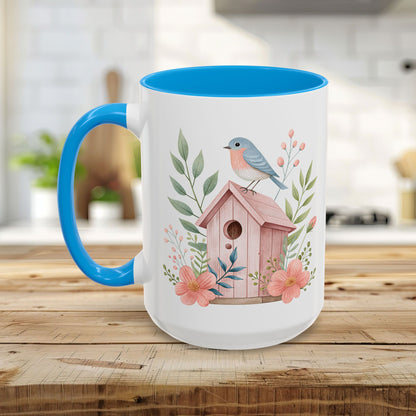 White ceramic mug with blue handle/interior, featuring a watercolor pink birdhouse, bluebird, and peach blossoms.