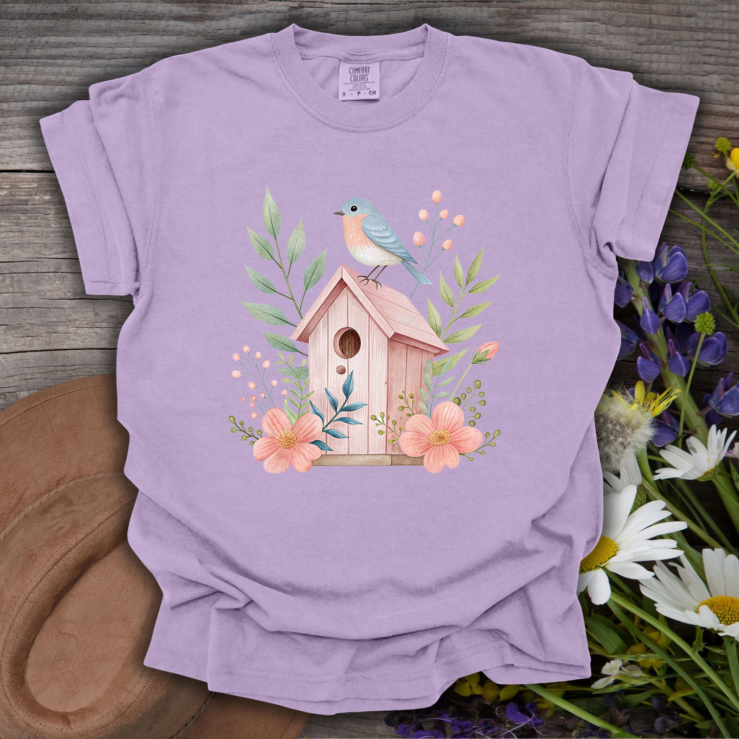 Orchid S t-shirt with a watercolor pink birdhouse, blue bird, and floral design on a wooden background.