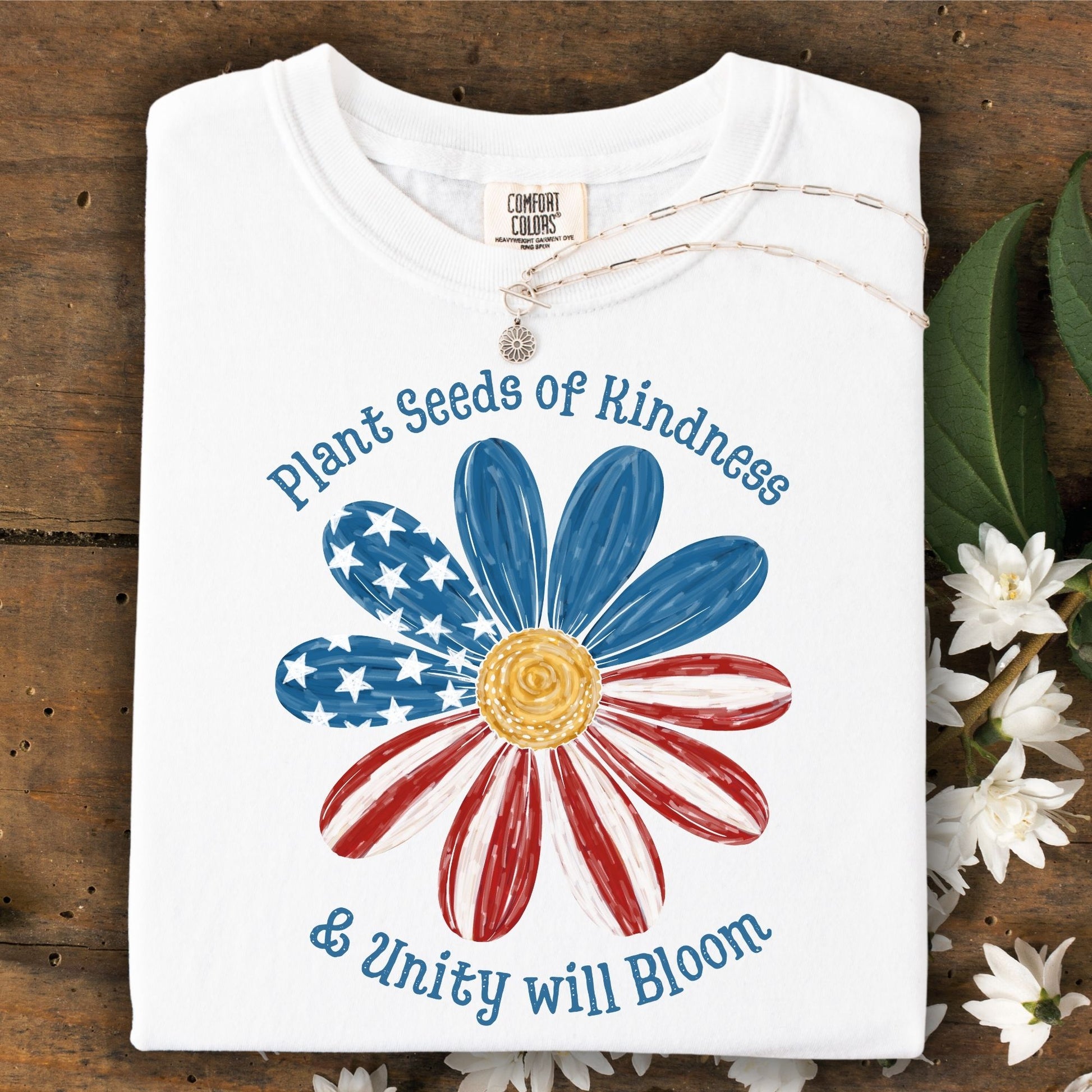White S-size t-shirt with "Plant Seeds of Kindness & Unity will Bloom" and a watercolor American flag daisy.