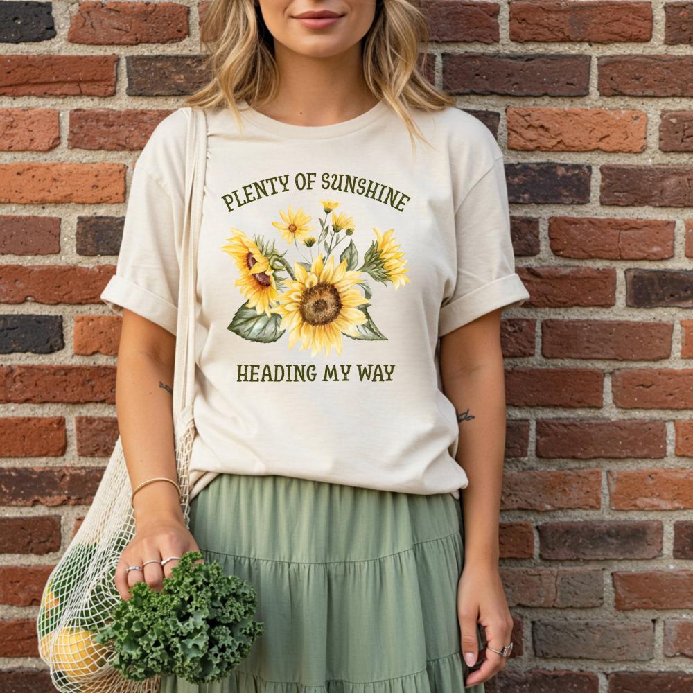 young woman wearing a t-shirt with sunflower design and text that reads "Plenty of sunshine heading my way" against a brick wall