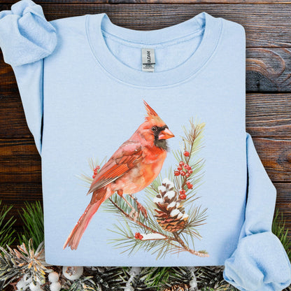 Light blue sweatshirt with a red cardinal on a snowy evergreen branch, pinecone, and berries.
