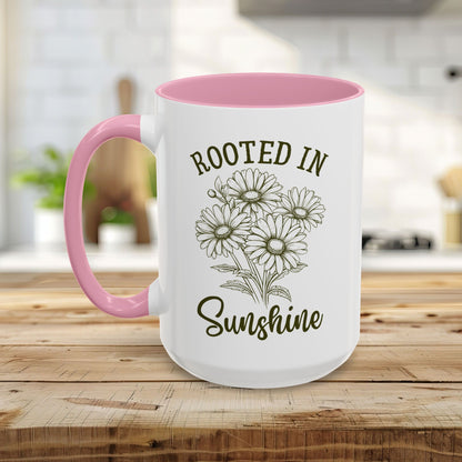 White ceramic mug with pink interior and handle, featuring "Rooted in Sunshine" and hand-drawn daisies.