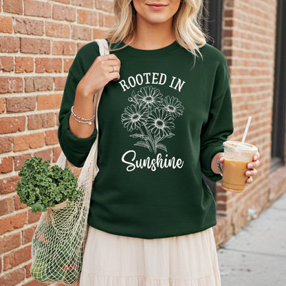 Woman in a dark green sweatshirt with "Rooted in Sunshine" and daisy graphic, holding coffee and a mesh bag of kale.