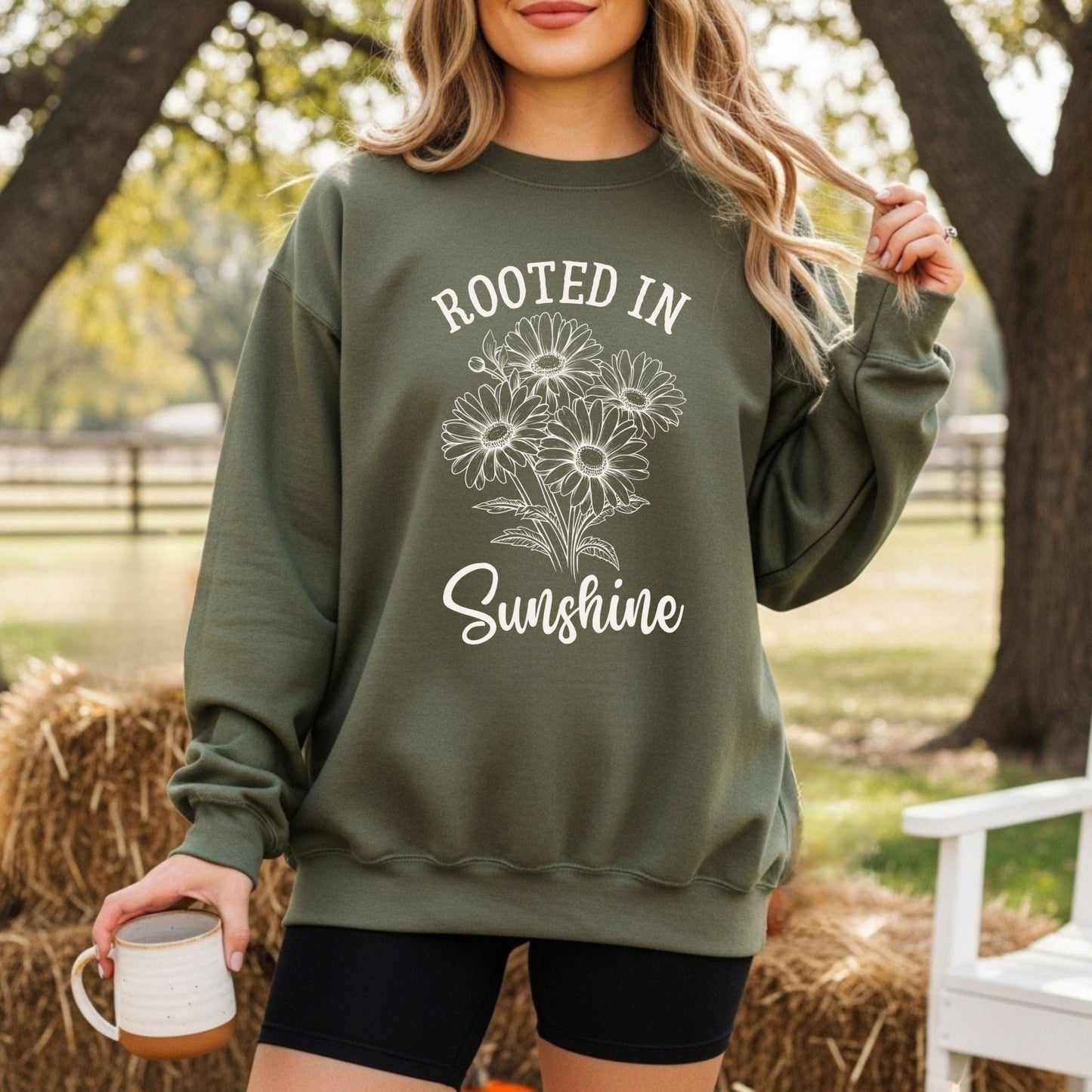 Woman wearing a forest green "Rooted in Sunshine" daisy sweatshirt, holding a mug outdoors.