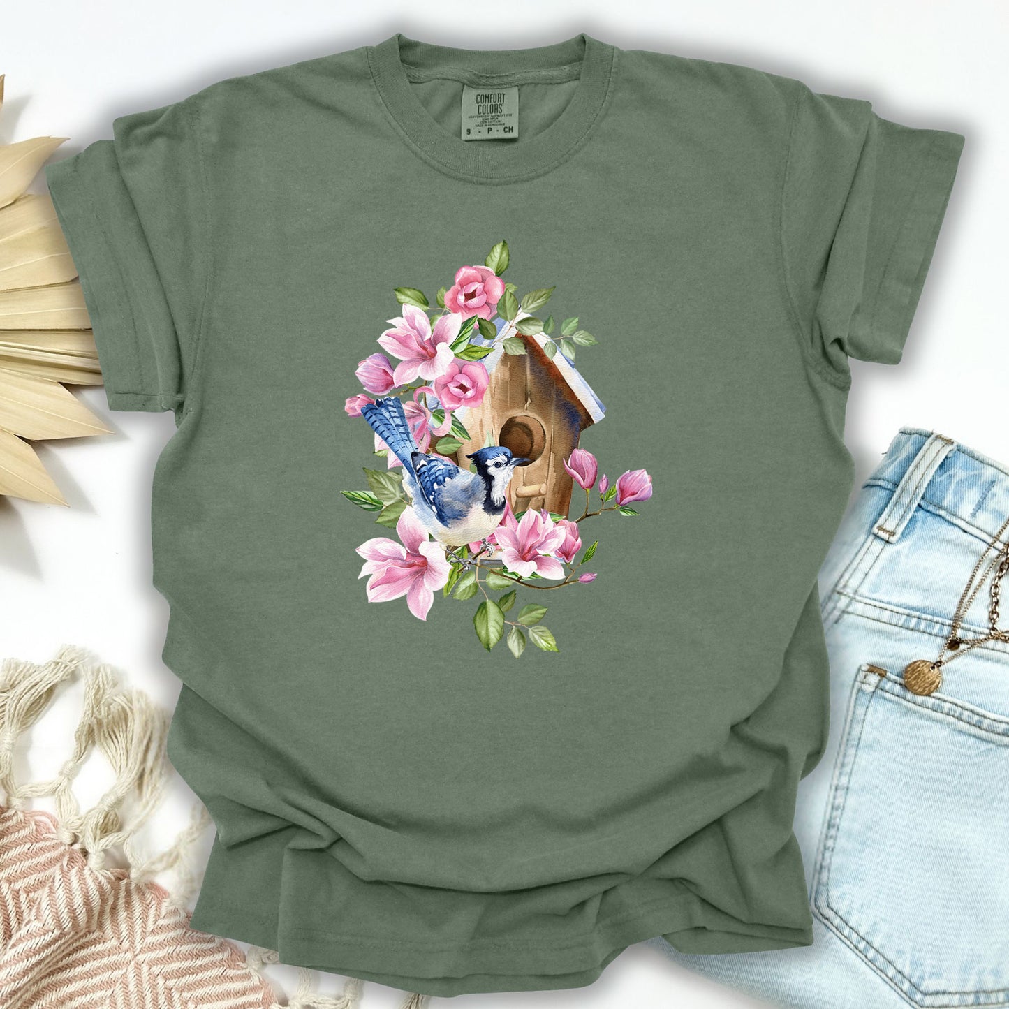 Moss S t-shirt with a watercolor blue jay on a rustic birdhouse surrounded by pink flowers.