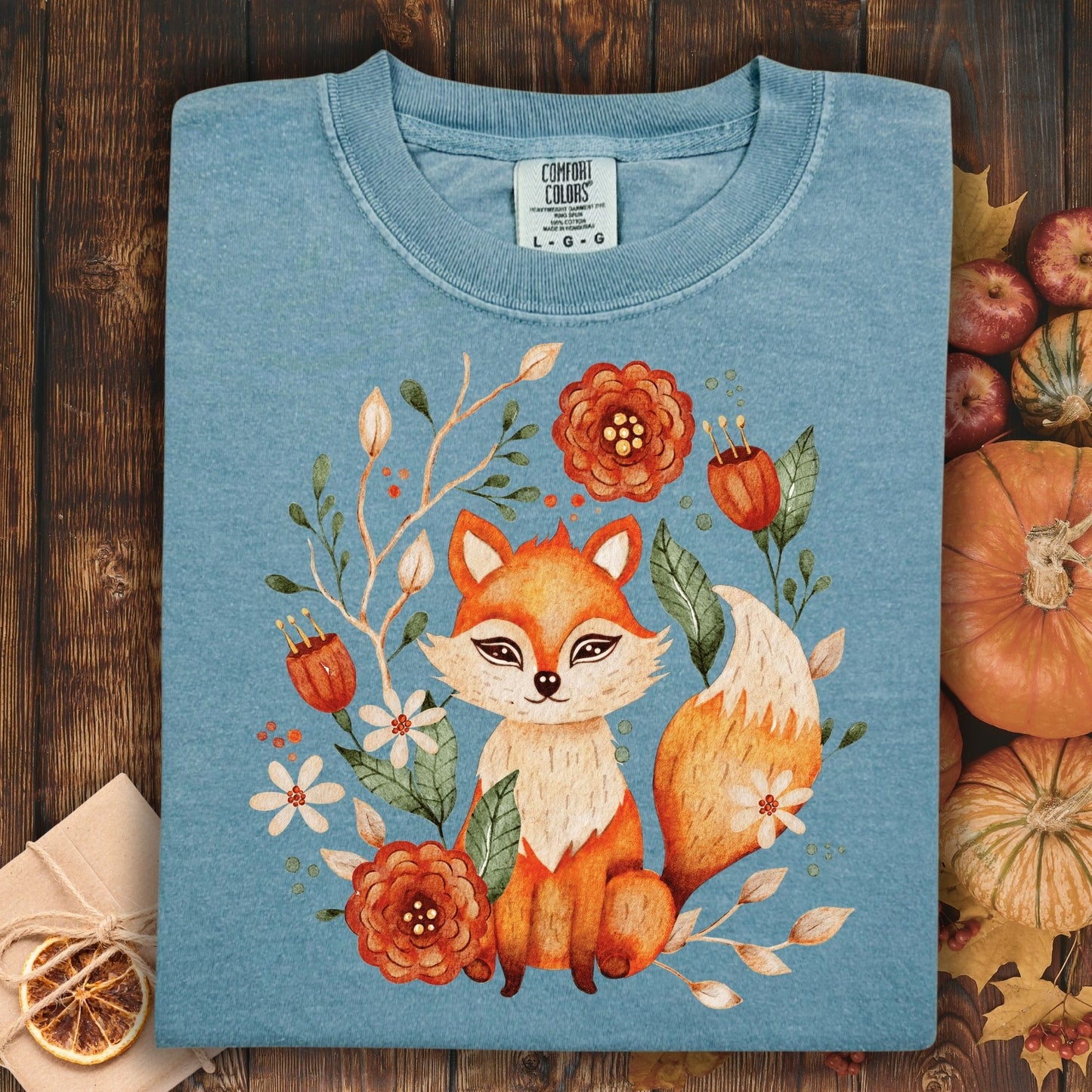 Folded Ice Blue t-shirt with a rustic folk art fox and floral design on a wooden background with autumn decor.