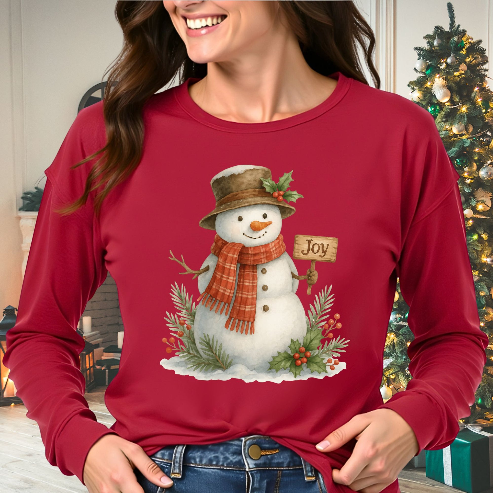 Woman in red long-sleeve tee with watercolor snowman holding "Joy" sign, holly, and snowy branches.