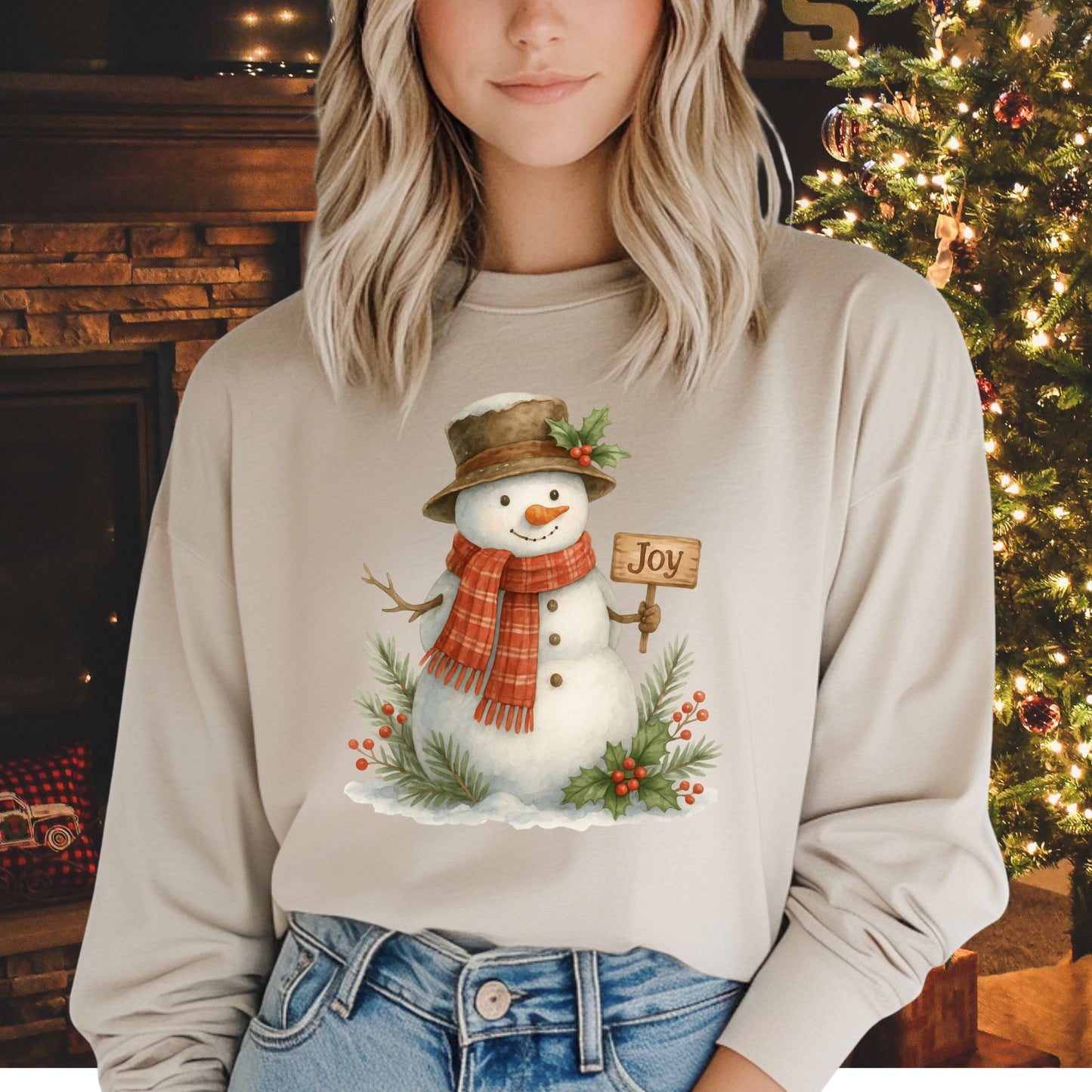Beige long-sleeve cotton tee with a watercolor snowman holding a "Joy" sign, holly, and snowy branches.