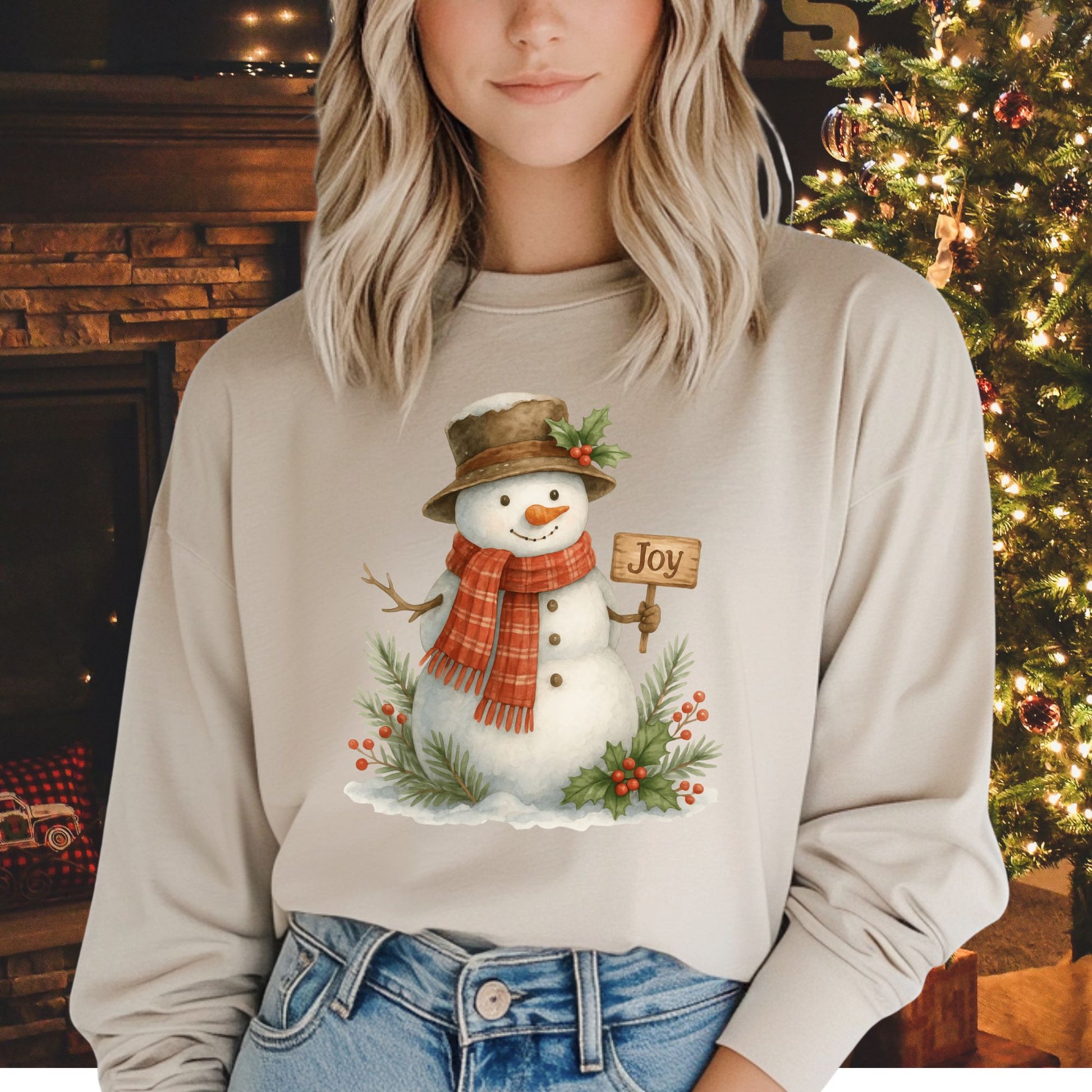 Beige long-sleeve cotton tee with a watercolor snowman holding a "Joy" sign, holly, and snowy branches.