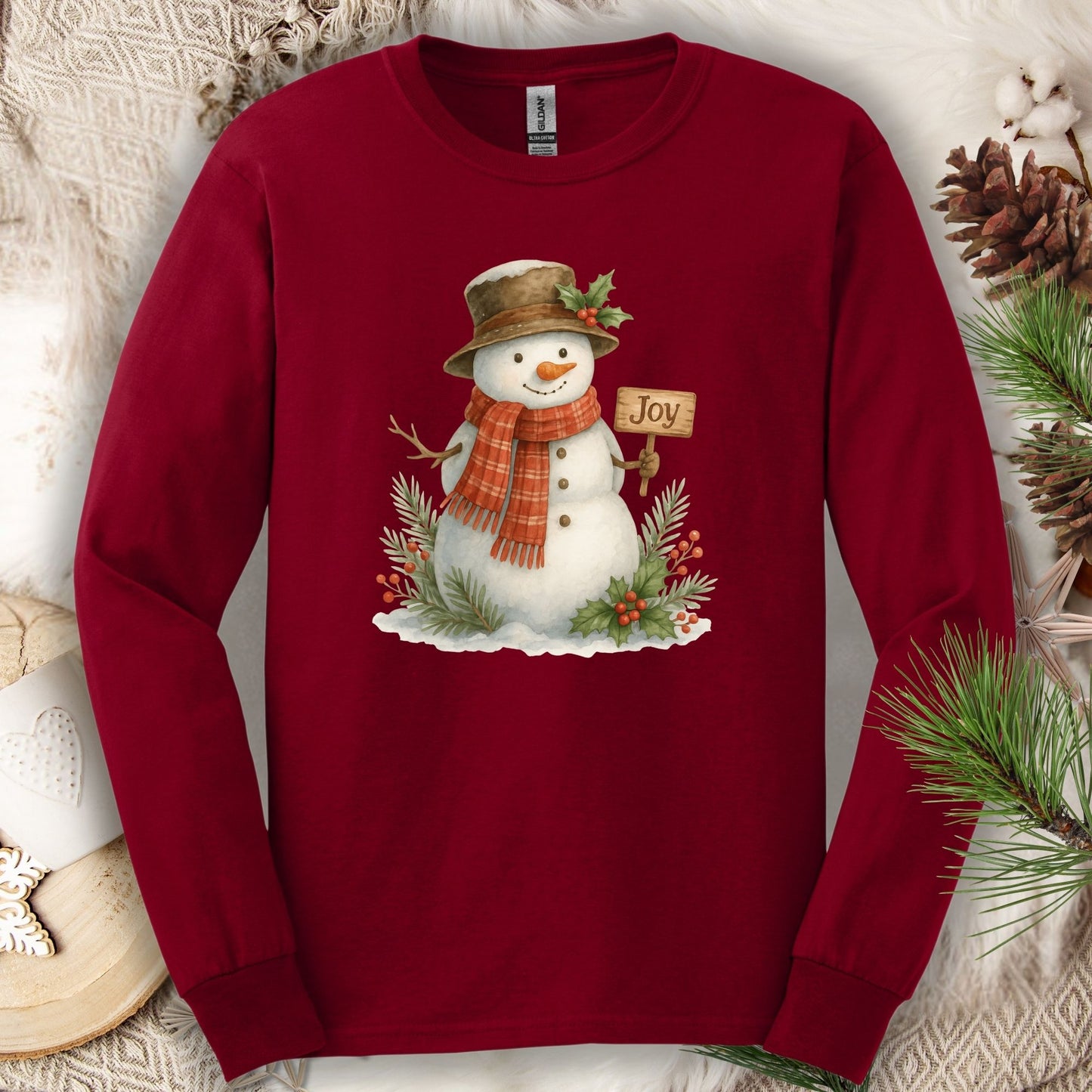 Cardinal Red long-sleeve tee with watercolor snowman holding a "Joy" sign, holly, and pine.