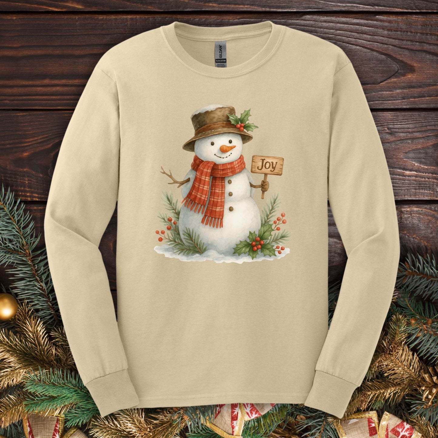 Sand long sleeve tee with a watercolor snowman holding a "Joy" sign, holly, and snowy branches.