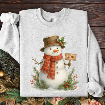 Ash Gildan 18000 sweatshirt with a watercolor snowman holding a "Joy" sign, surrounded by holly and snowy branches.