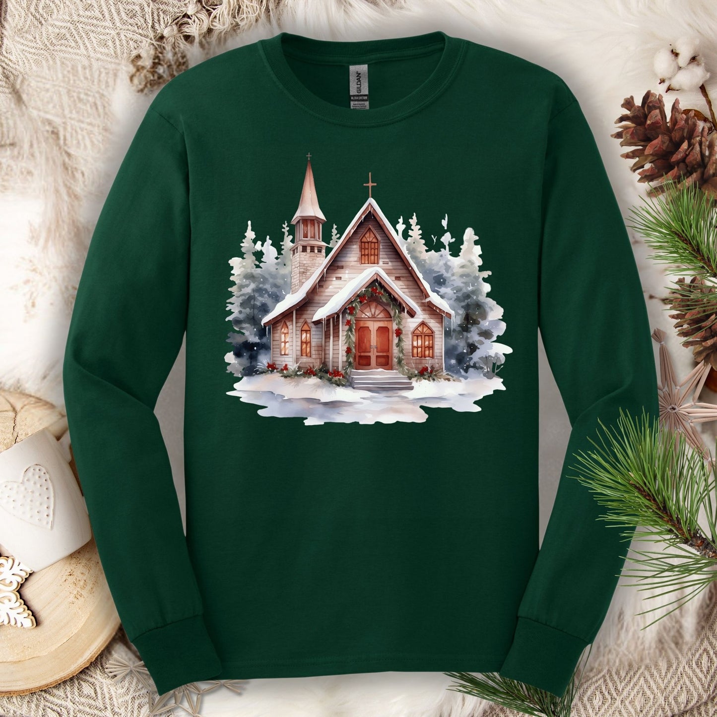 Forest Green Gildan long sleeve tee with a rustic wooden chapel in snow, decorated with holly.