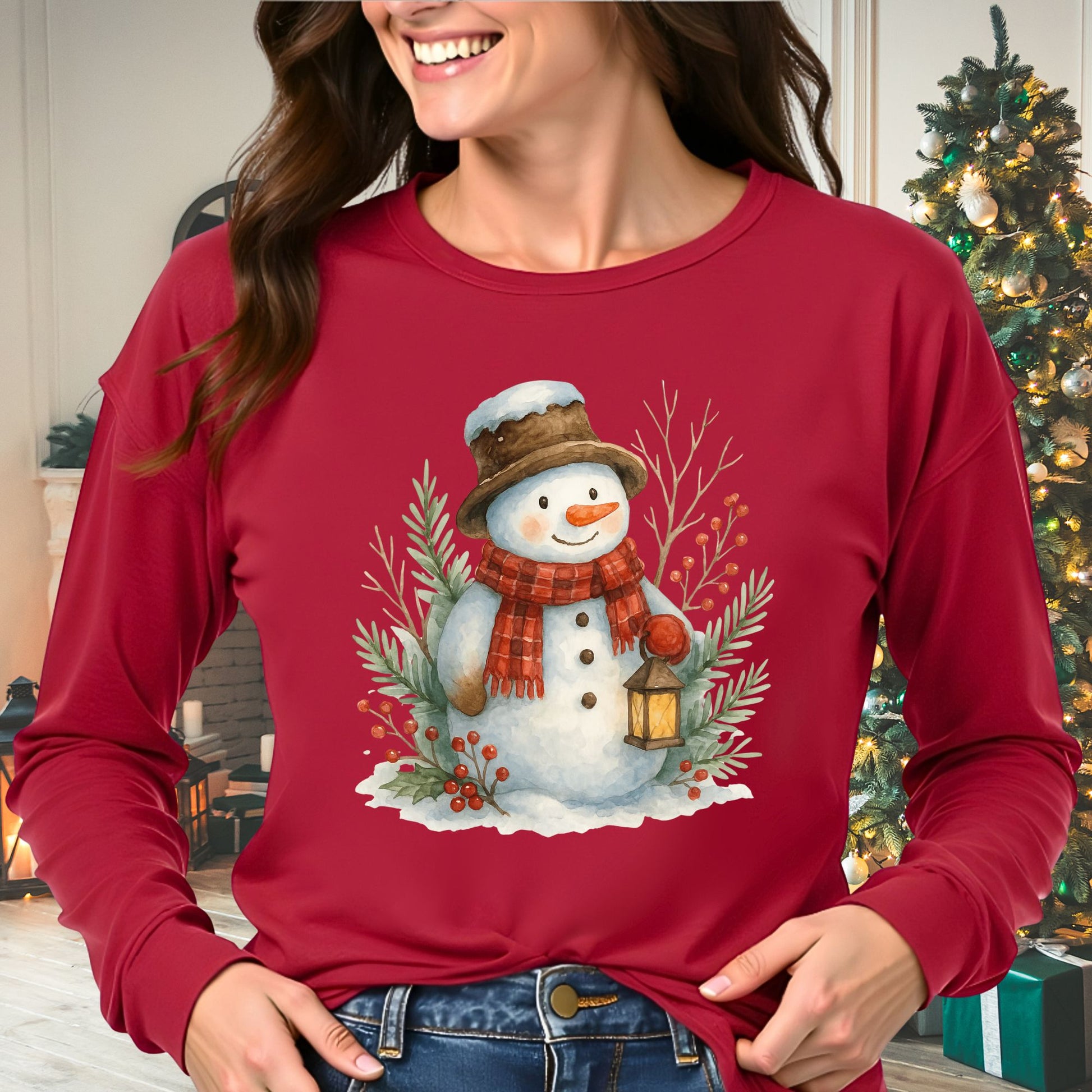 Woman in a red long-sleeve tee with a watercolor snowman holding a lantern, surrounded by snowy branches.