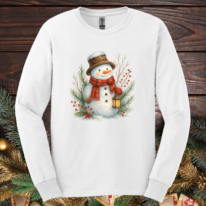 White long-sleeve tee with a watercolor snowman holding a lantern, surrounded by snowy branches.