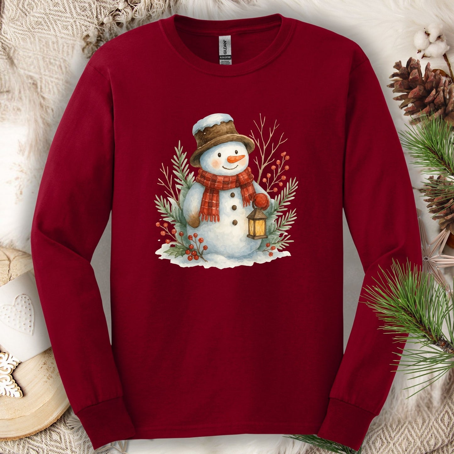 Cardinal Red long sleeve tee with a watercolor snowman holding a lantern, surrounded by snowy branches.