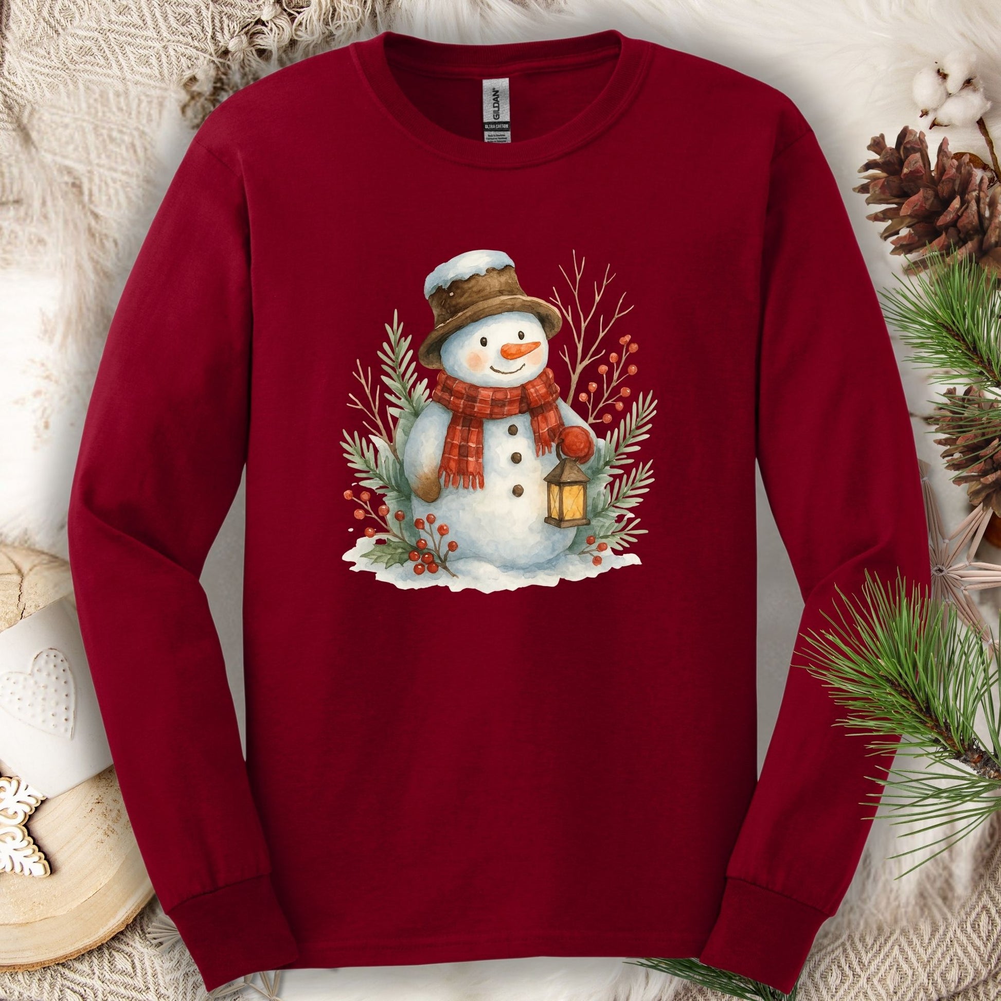 Cardinal Red long sleeve tee with a watercolor snowman holding a lantern, surrounded by snowy branches.