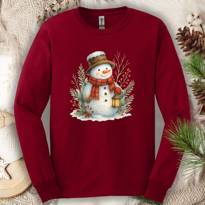 Cardinal Red long sleeve tee with a watercolor snowman holding a lantern, surrounded by snowy branches.