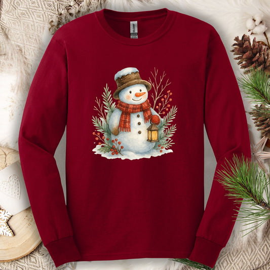 Cardinal Red long sleeve tee with a watercolor snowman holding a lantern, surrounded by snowy branches.