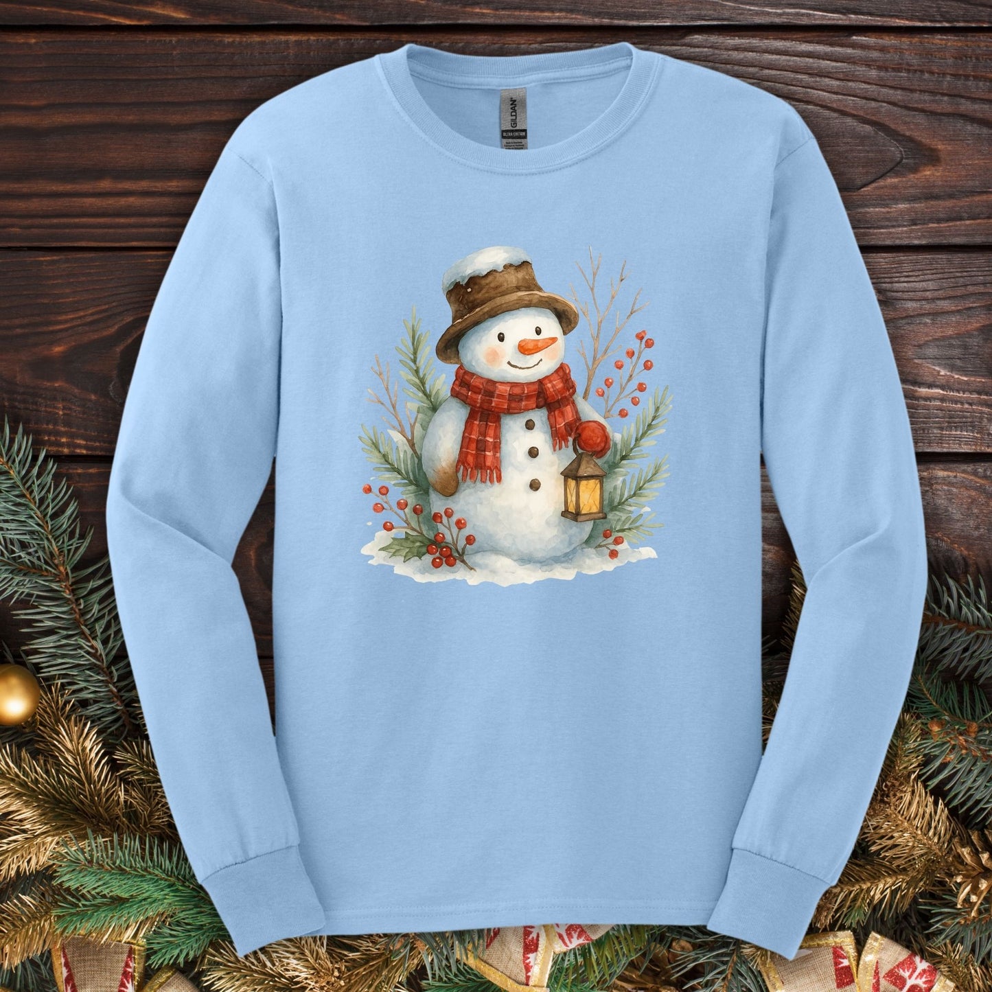 Light blue Gildan long sleeve tee with a watercolor snowman holding a lantern, surrounded by snowy branches.
