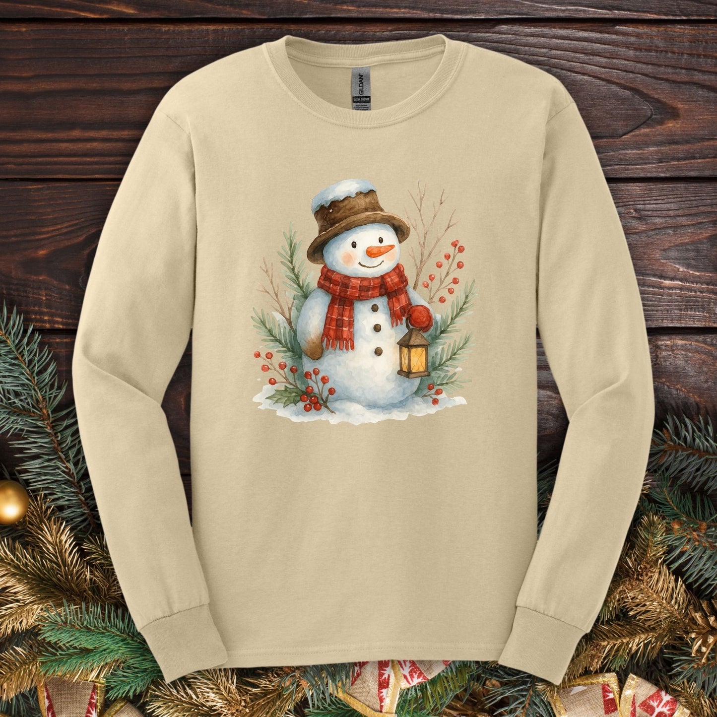 Sand long sleeve tee with a watercolor snowman holding a lantern, surrounded by snowy branches.