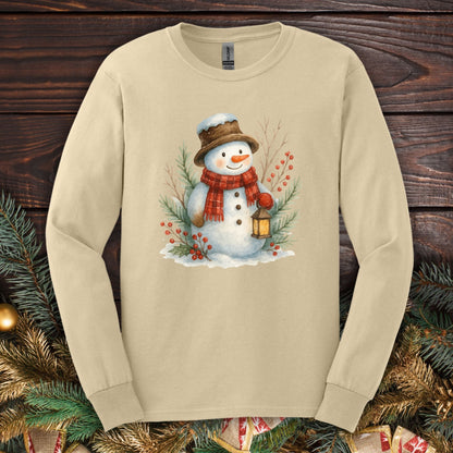 Sand long sleeve tee with a watercolor snowman holding a lantern, surrounded by snowy branches.