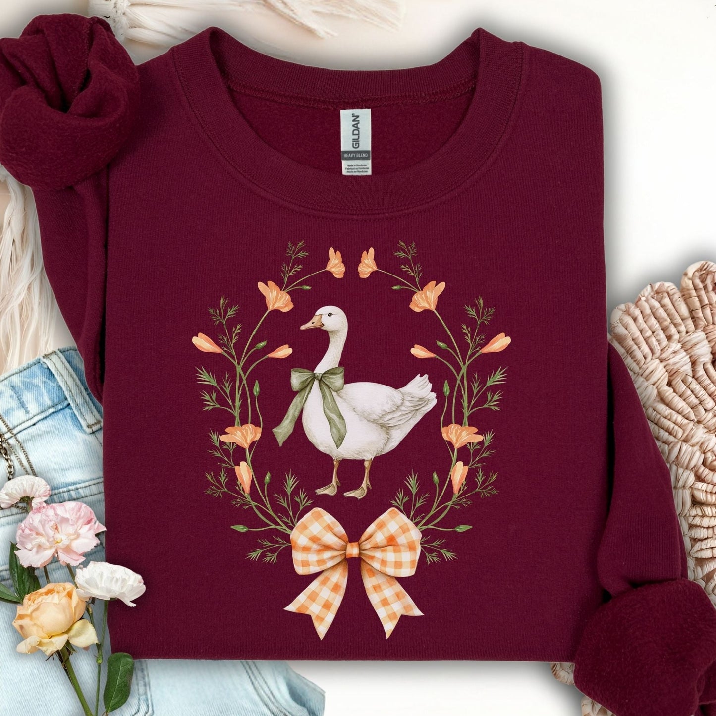 Maroon crewneck sweatshirt with a white goose in a floral wreath, tied with a green ribbon and orange gingham bow.