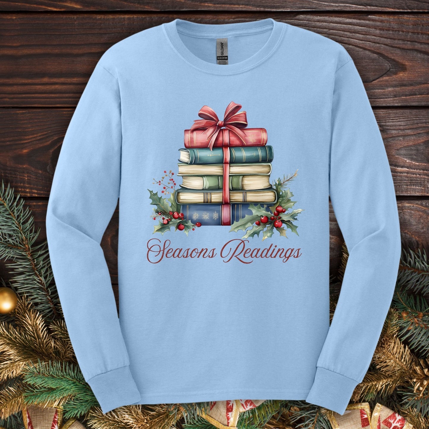Light blue long sleeve tee with watercolor stacked Christmas books, red bow, holly, and "Seasons Readings" text.