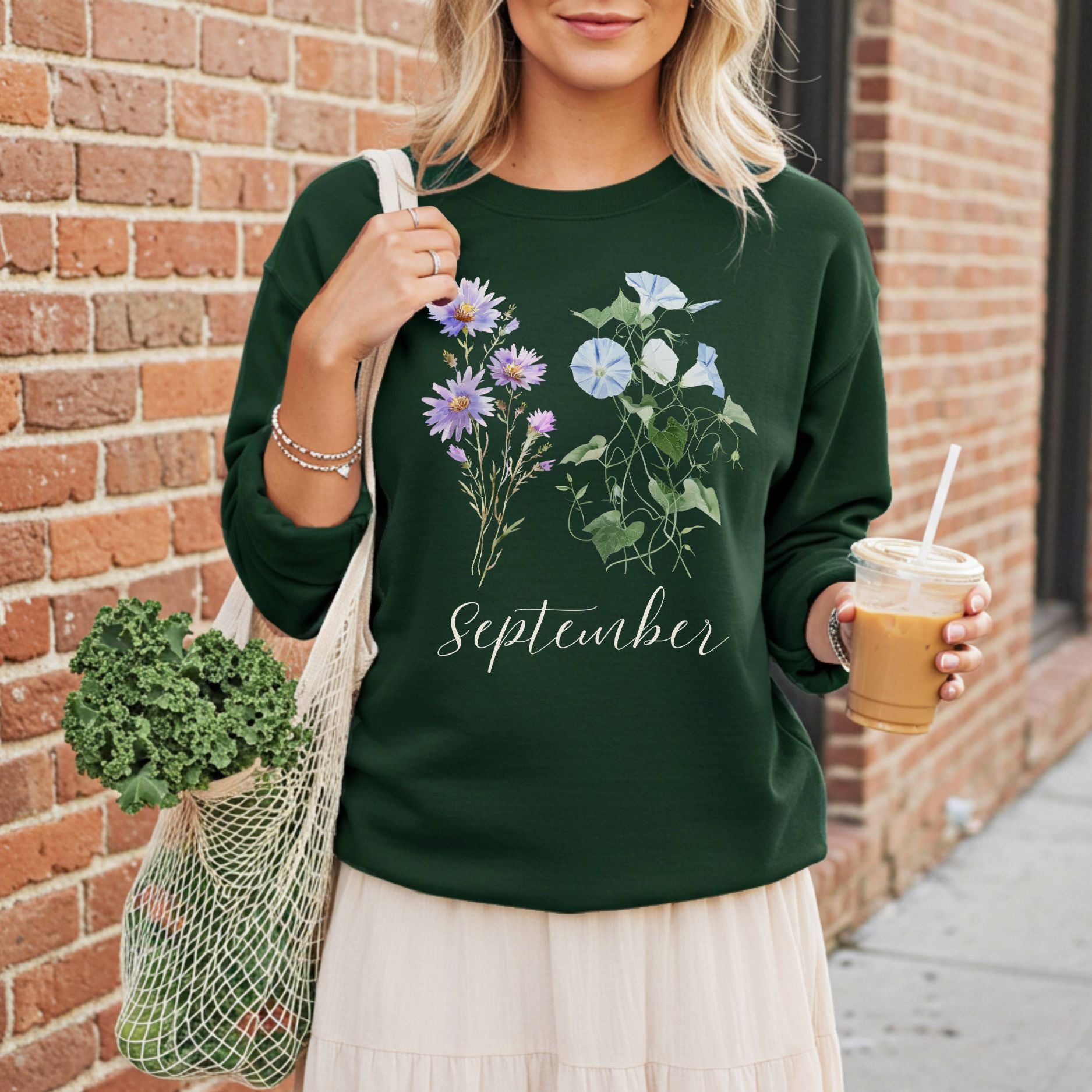 Woman in a dark green crewneck sweatshirt with watercolor morning glory and aster flowers, holding a coffee and market bag.