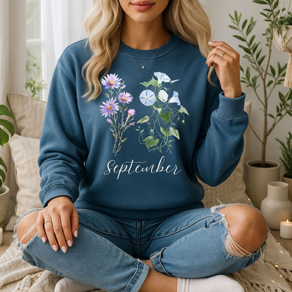 Woman in a blue crewneck sweatshirt with watercolor morning glory and aster flowers and "September" text.