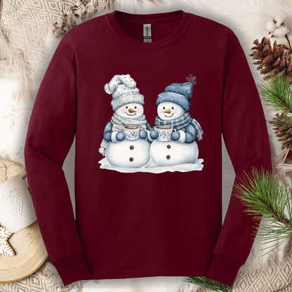 Maroon long-sleeve tee with two cozy snowmen holding hot chocolate mugs, surrounded by winter decor.