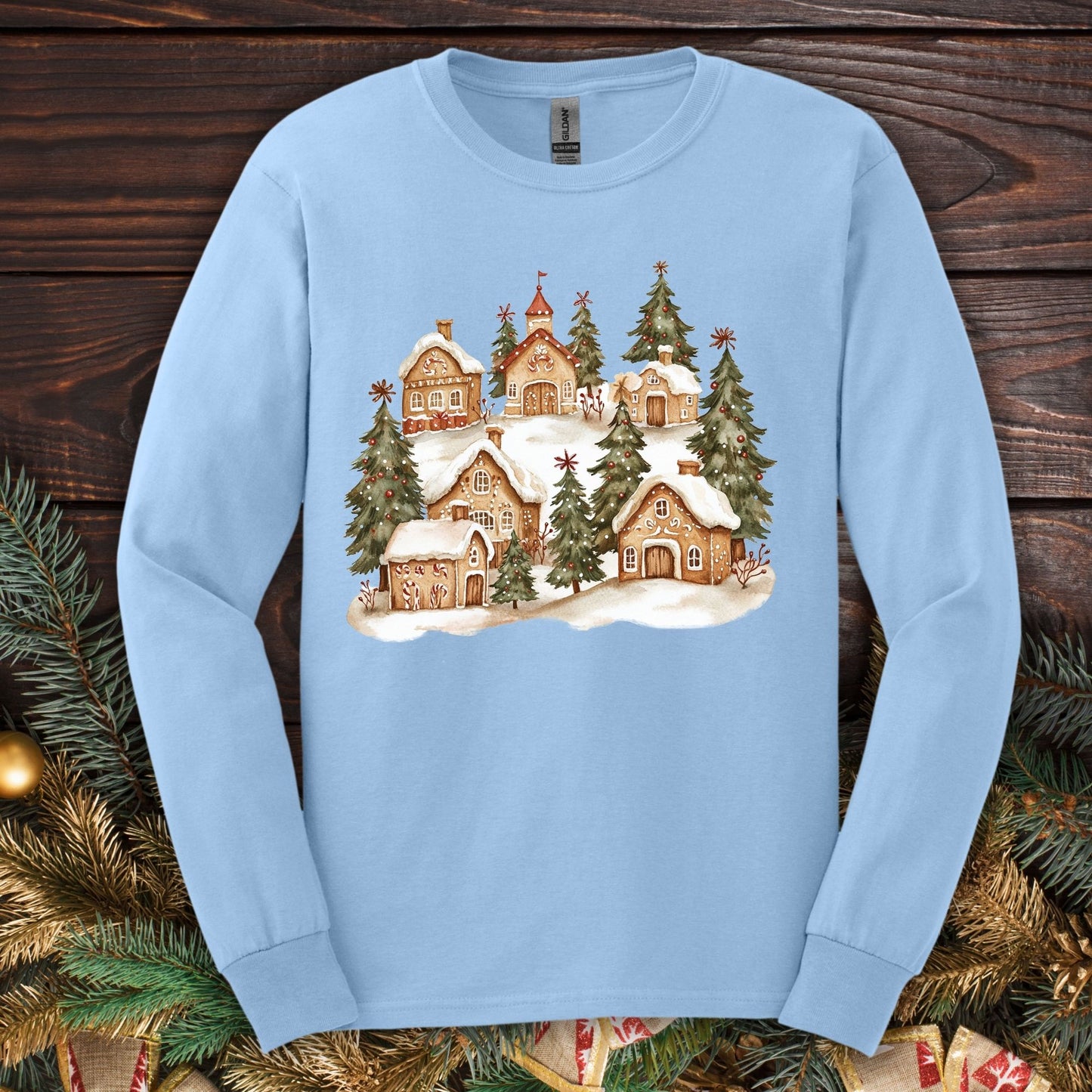 Light blue long sleeve tee with a watercolor snowy gingerbread village and evergreens.