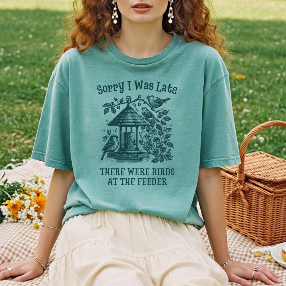 young woman wearing a teal t-shirt with a bird-themed design that reads "Sorry I was Late. There were birds at the feeder" in a park setting.