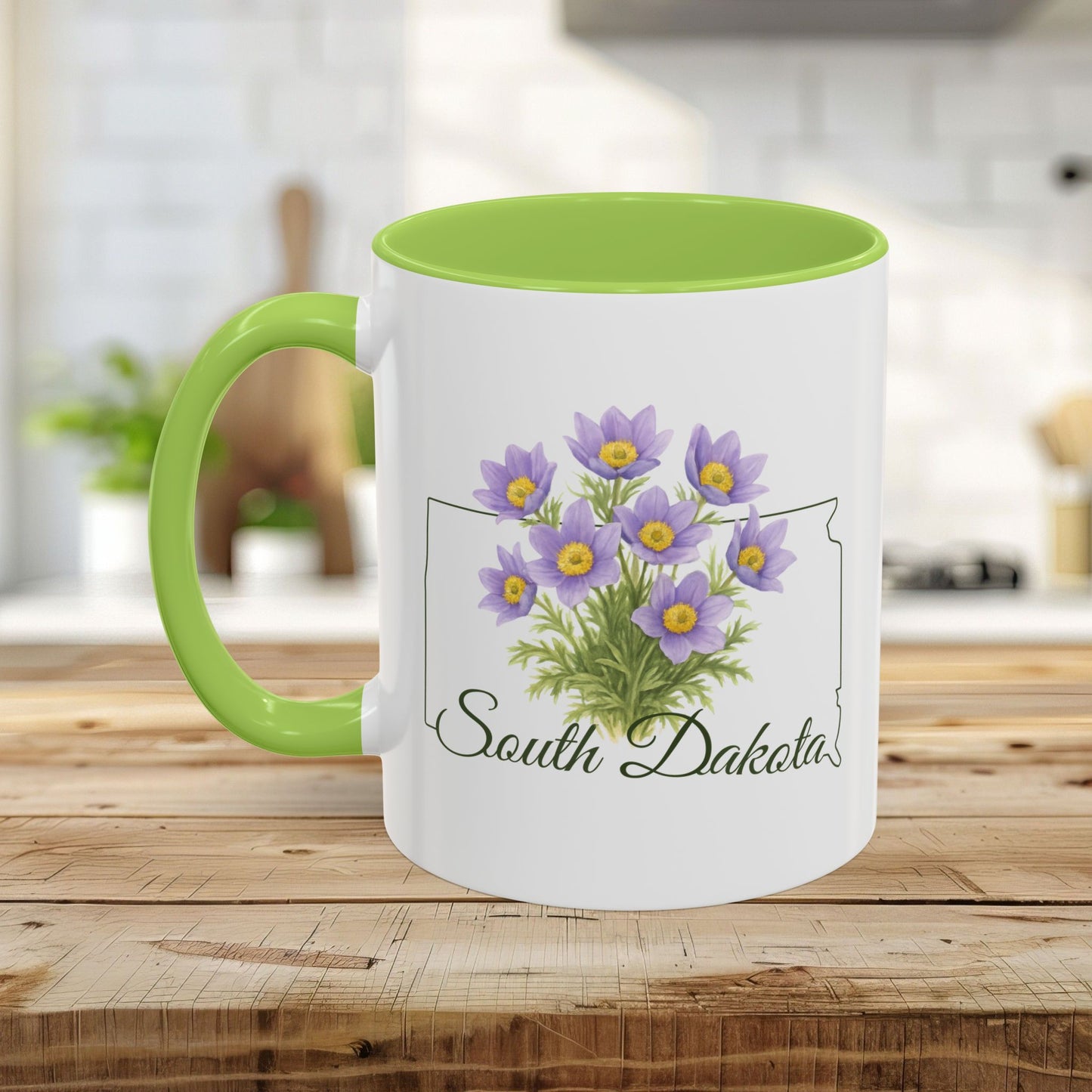 White ceramic mug with green handle and interior, featuring a watercolor American Pasque flower bouquet and "South Dakota" script over a state outline.