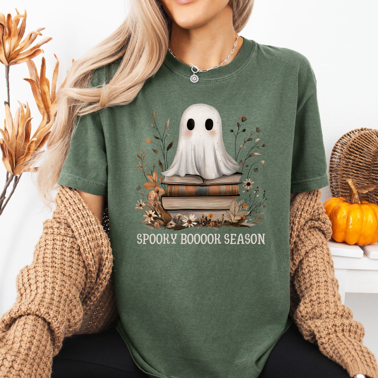 Green tee with a cute ghost on vintage books, "Spooky Boooook Season" text, surrounded by fall foliage.