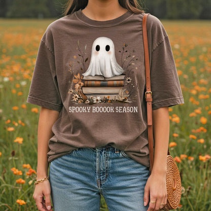 Brown "Spooky Booook Season" tee with a ghost on vintage books, worn by a person in a field.