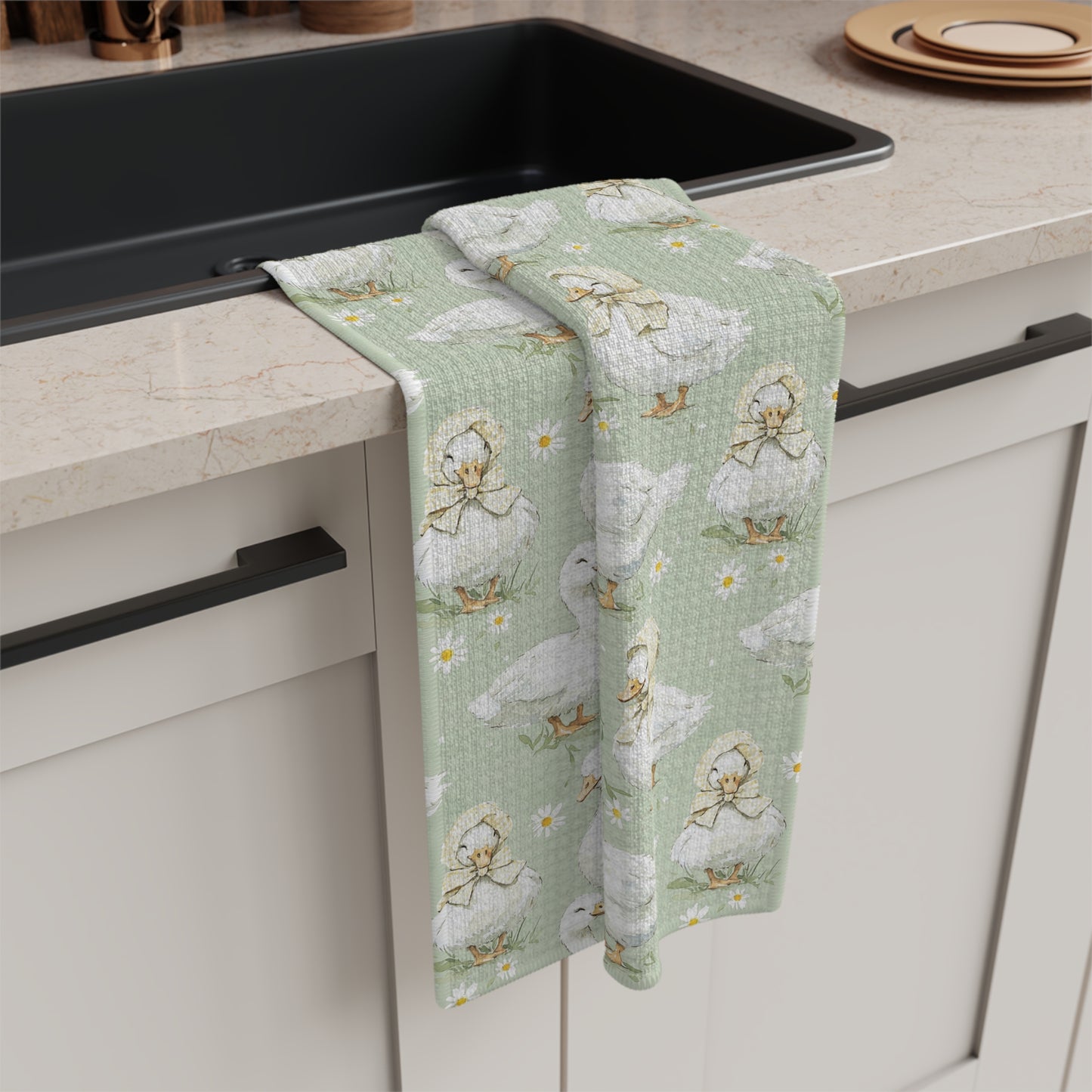 Microfiber tea towel with white ducks and ducklings in bonnets on a sage background, draped over a kitchen sink.