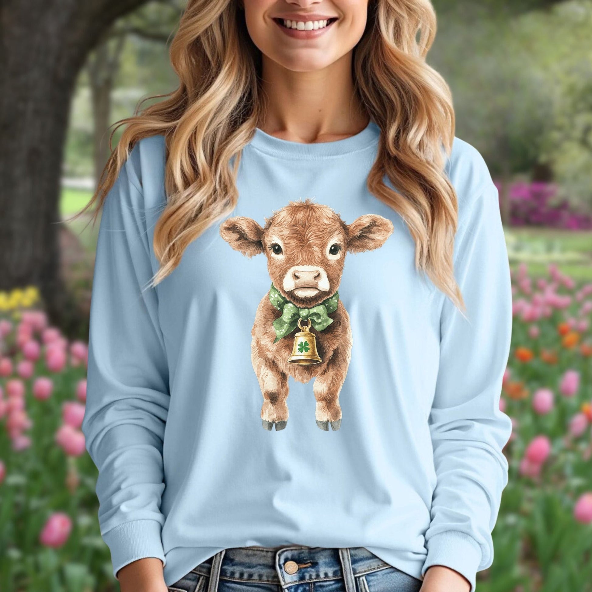 Light blue long-sleeve tee with a watercolor baby Highland cow wearing a green bow and clover bell.