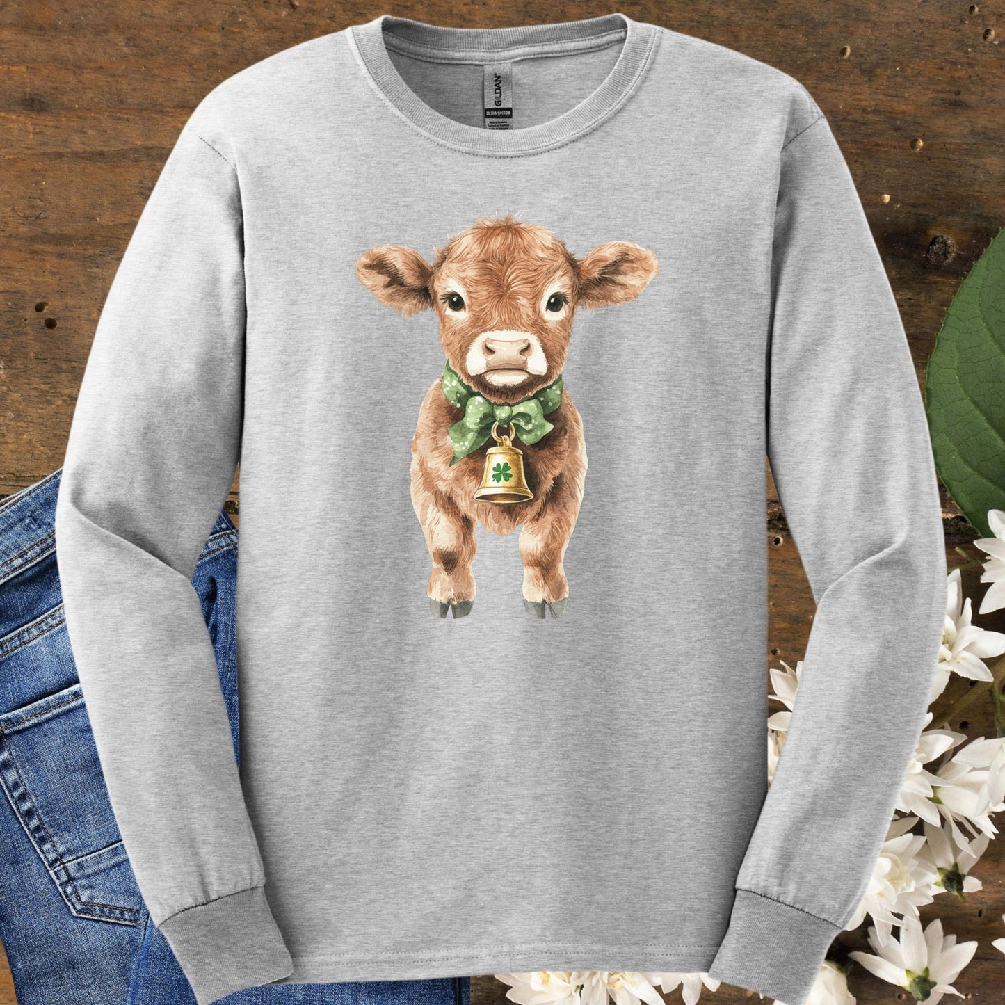 Ash long-sleeve tee with a watercolor baby Highland cow wearing a green bow and clover bell.