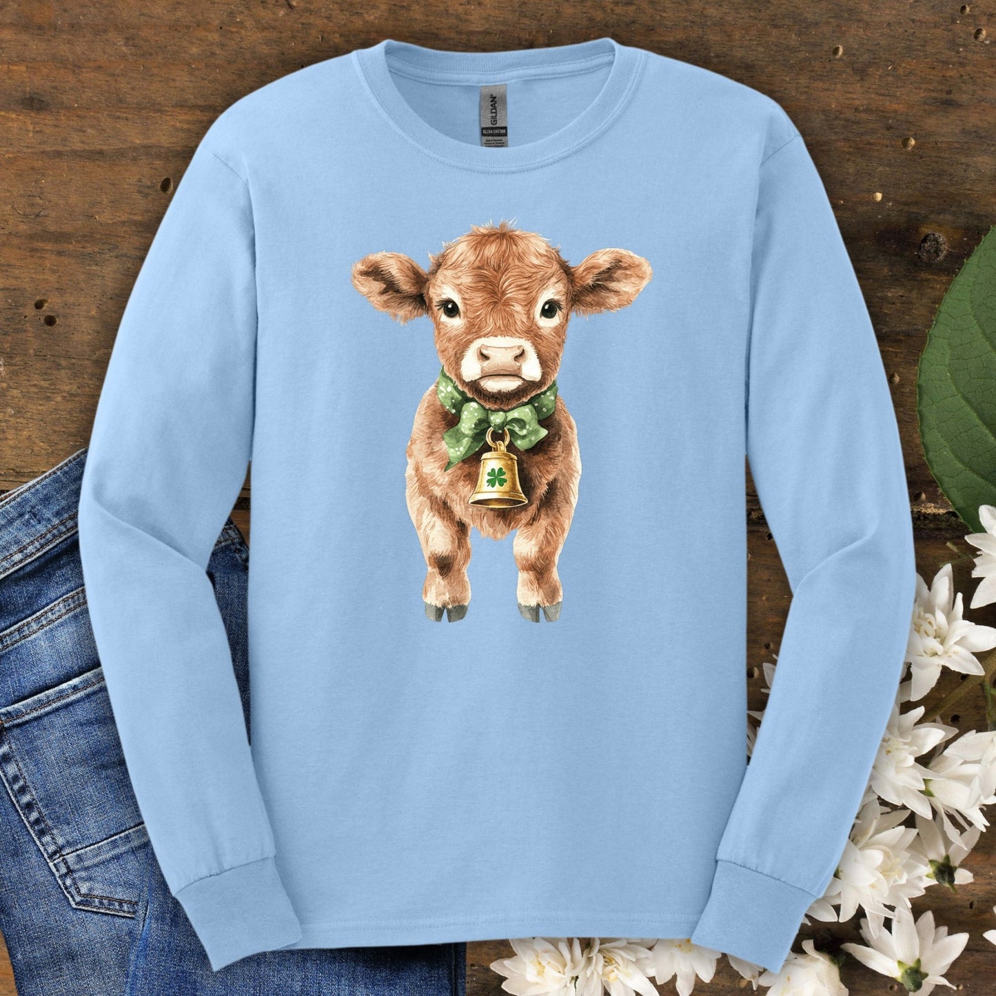 Light blue long sleeve tee with a watercolor baby Highland cow wearing a green bow and clover bell.