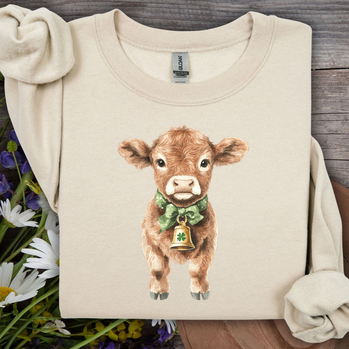 Sand unisex sweatshirt with watercolor baby Highland cow, green bow, and clover bell.
