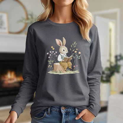 Woman in a dark gray long-sleeve tee with a whimsical rabbit reading on a tree stump surrounded by flowers.
