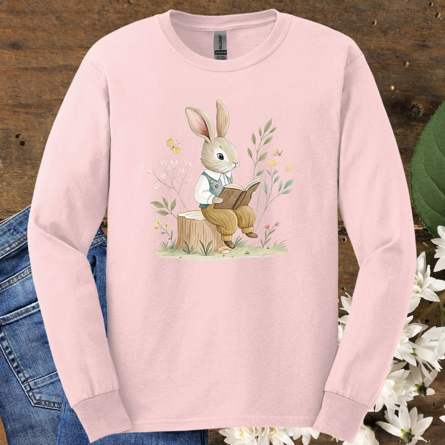 Light pink long-sleeve tee with a watercolor rabbit reading on a stump, surrounded by botanicals.