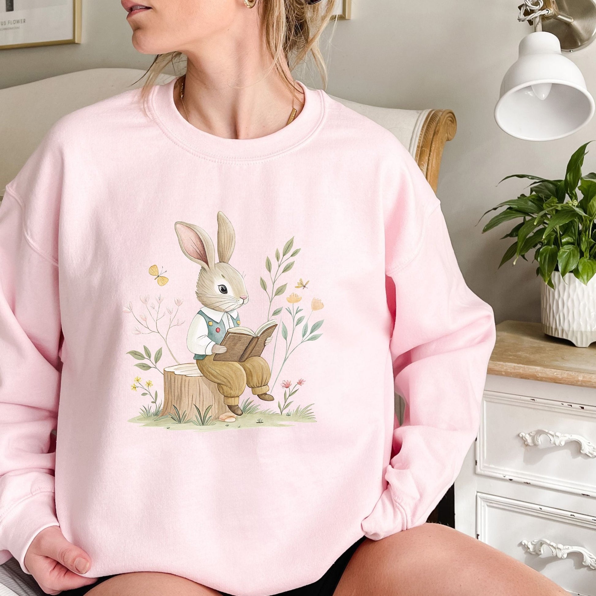 Pink sweatshirt with a watercolor illustration of a bunny reading on a tree stump in a garden.