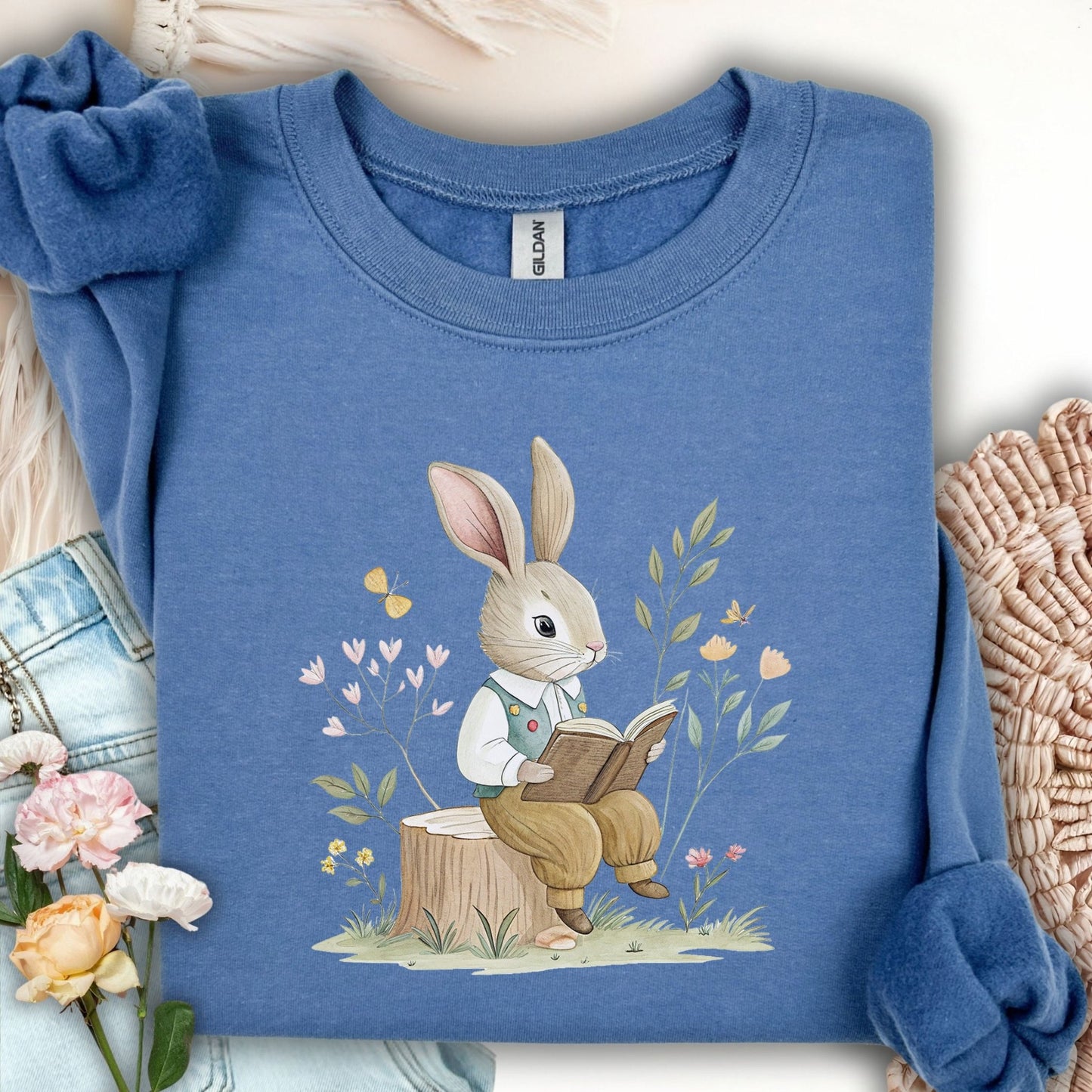 Indigo Blue sweatshirt with a charming rabbit reading on a tree stump, surrounded by spring flowers and butterflies.