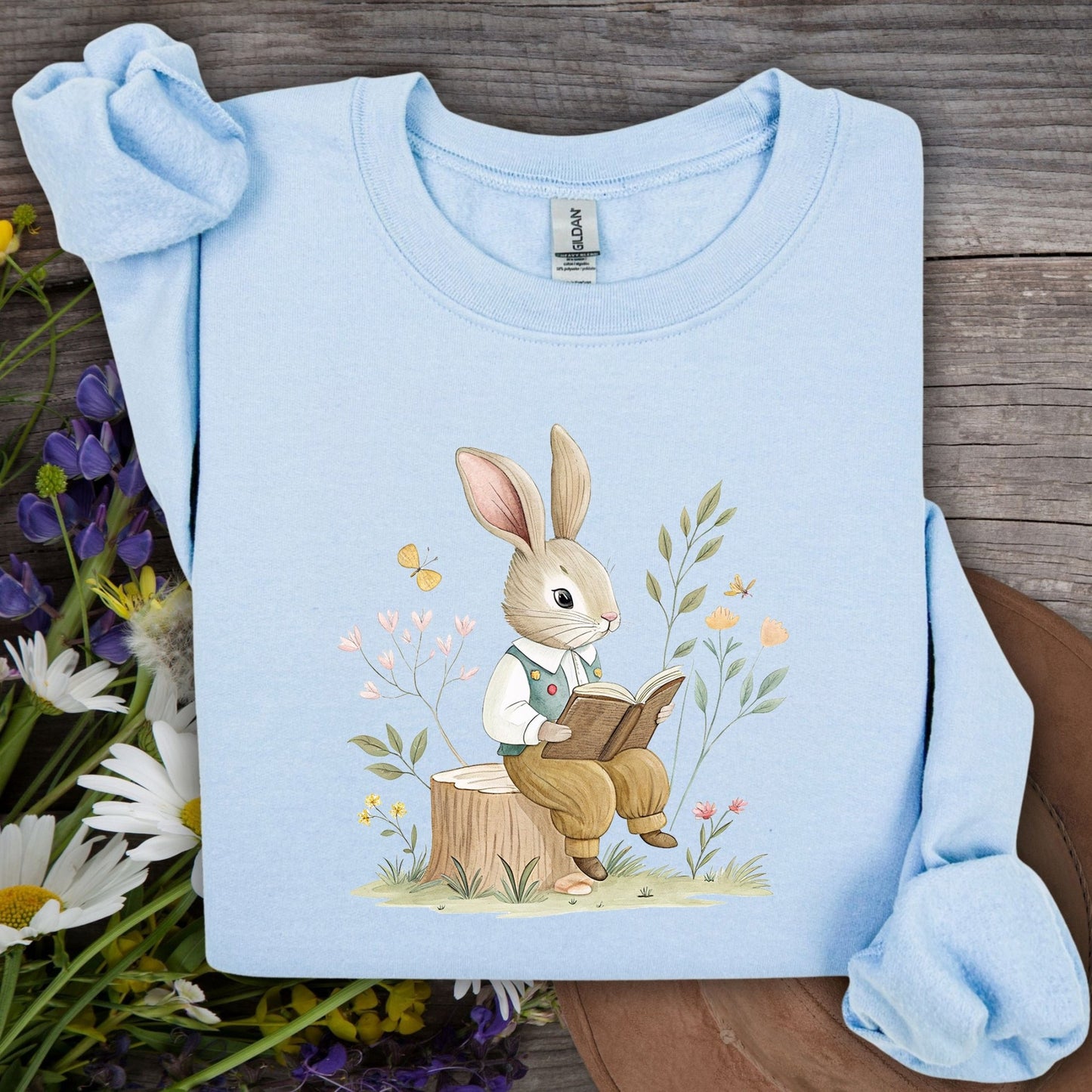 Light blue sweatshirt with a bookish bunny in a waistcoat reading on a tree stump, surrounded by spring flowers.